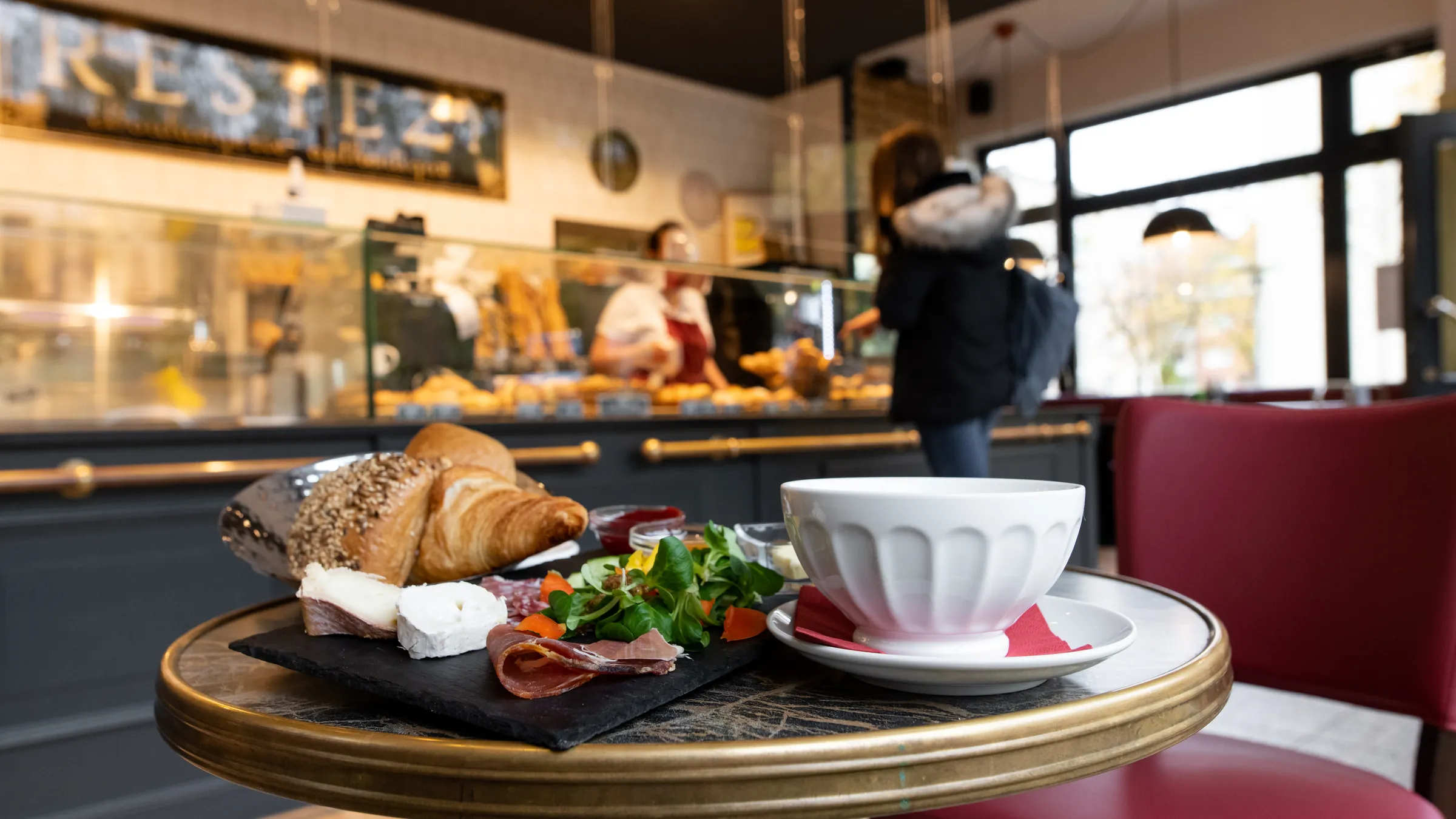 Frühstücksteller und Café au lait auf einem Bistrotisch im à table, dahinter die Theke